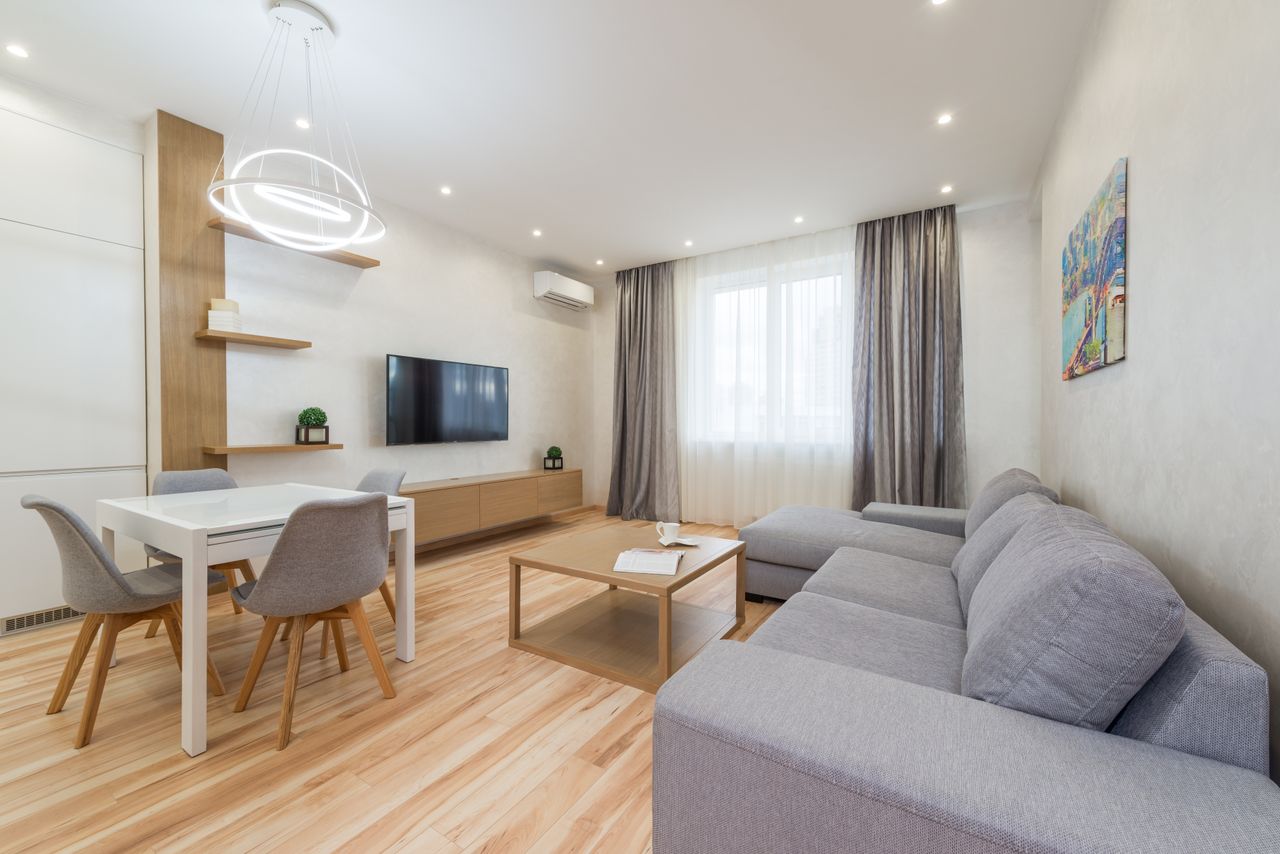 Interior of modern bright living room with couch near table and chairs next to TV on wall and shelves near curtains on windows